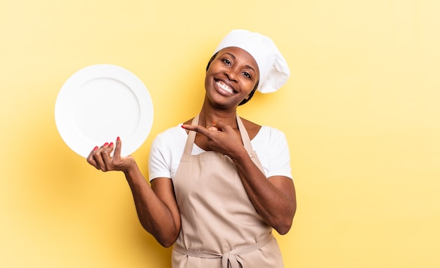 Mulher negra chef afro sorrindo alegremente, sentindo-se feliz e apontando para o lado e para cima, mostrando o objeto no espaço da cópia. conceito de prato vazio