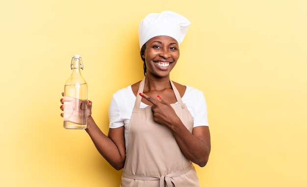 Mulher negra chef afro sorrindo alegremente, sentindo-se feliz e apontando para o lado e para cima, mostrando o objeto no espaço da cópia segurando uma garrafa de água