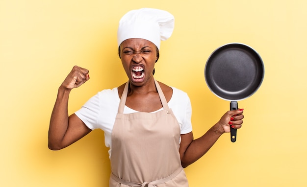 Mulher negra chef gritando agressivamente com uma expressão de raiva ou com os punhos cerrados celebrando o sucesso