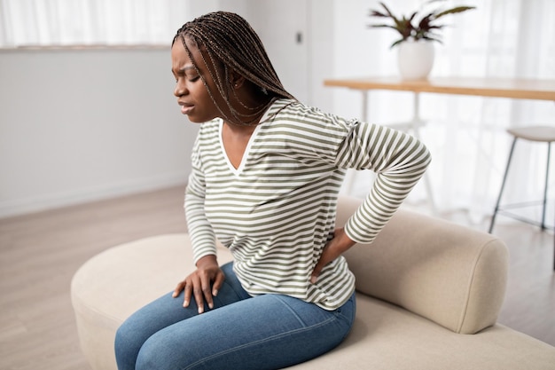 Mulher negra com dor sentada no sofá tocando a parte inferior das costas