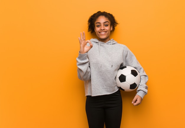 Mulher negra de aptidão jovem alegre e confiante fazendo o gesto de ok. Segurando uma bola de futebol.