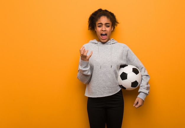 Mulher negra de aptidão jovem muito assustada e com medo. Segurando uma bola de futebol.