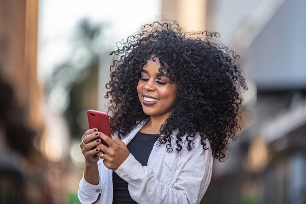 Mulher negra de cabelo encaracolado jovem andando usando telefone celular. Mensagens de texto na rua. Cidade grande.