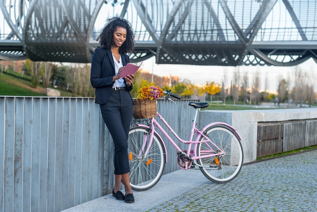 Mulher negra de negócios com bicicleta vintage pelo rio, olhando para a agenda dela