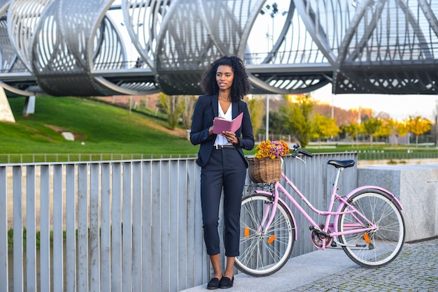 Mulher negra de negócios com bicicleta vintage pelo rio, olhando para a agenda dela