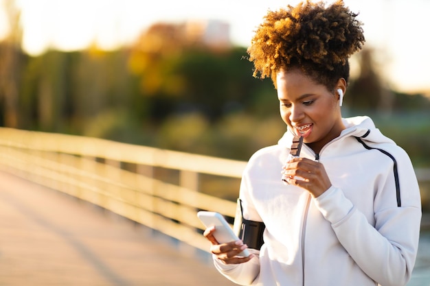 Mulher negra descansando após treinamento ao ar livre comendo barra de proteína e usando smartphone