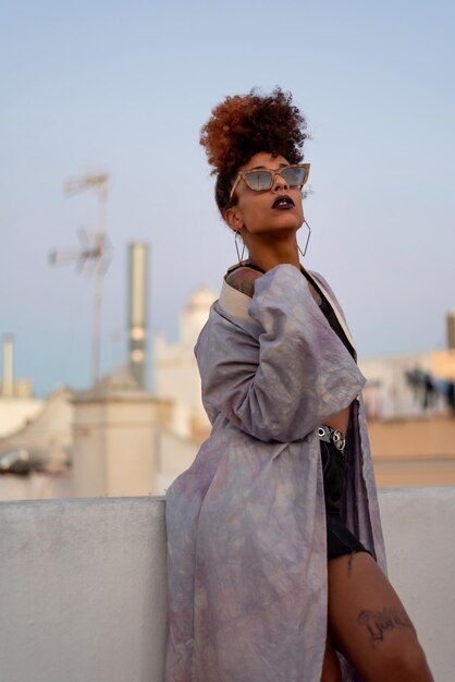 Mulher negra elegante com cabelo encaracolado e óculos de sol vestindo um manto lilás enquanto descansava no terraço contra o céu azul sem nuvens em Cádiz Espanha