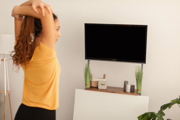 Mulher negra esticando os braços se exercitando olhando para a tela da televisão dentro de casa