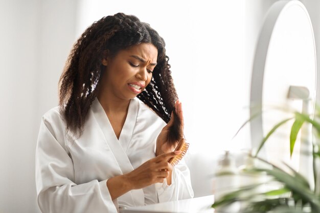 Foto mulher negra estressada penteando o cabelo emaranhado com escova de bambu em casa