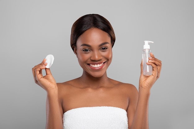 Mulher negra feliz segurando algodão e garrafa com gel de limpeza facial