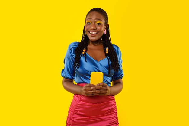 Mulher negra feliz usando telefone em pé no fundo amarelo do estúdio