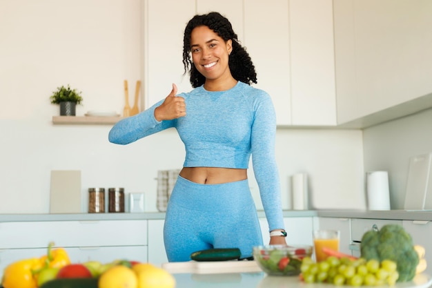 Mulher negra feliz vestindo roupas esportivas e aparecendo o polegar enquanto prepara salada saudável na cozinha