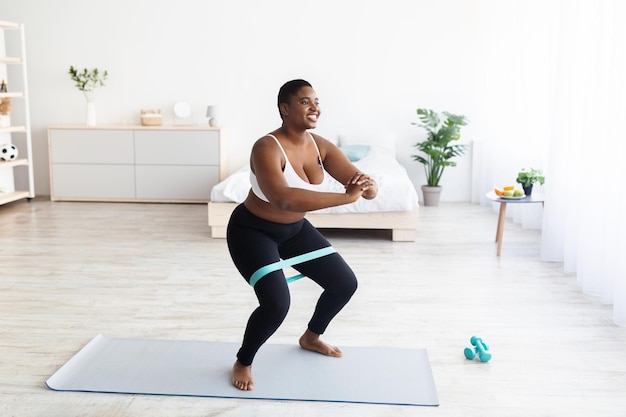 Foto mulher negra gordinha milenar de cócoras com elástico durante o espaço livre de treino doméstico