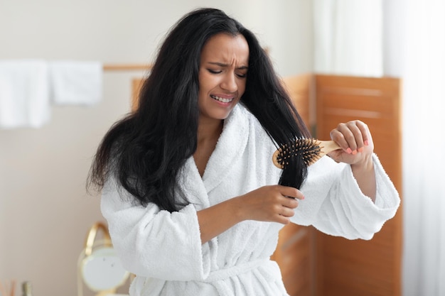 Mulher negra infeliz escovando cabelos desembaraçados com escova de cabelo no banheiro