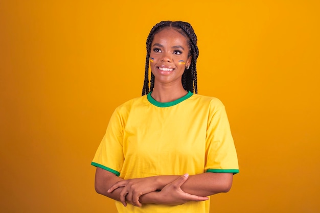 Mulher negra jovem fã de futebol brasileiro braços cruzados confiante feliz alegre Jovem afro brasileira líder de torcida com braços cruzados em fundo amarelo