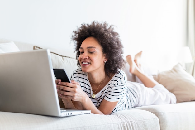 Mulher negra jovem feliz usando o celular enquanto está sentado em um sofá em casa com computador portátil.