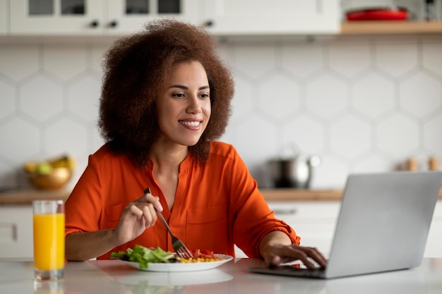 Mulher negra jovem sorridente usando computador portátil durante o café da manhã na cozinha