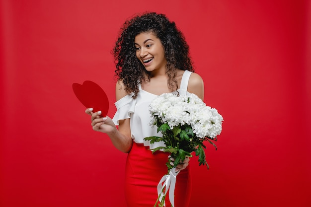 Foto mulher negra, lendo o coração em forma de cartão de dia dos namorados e buquê de flores brancas, isoladas no vermelho