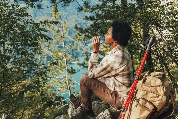 Mulher negra madura fazendo uma pausa na água e apreciando a vista no topo de uma colina durante caminhada nas montanhas.