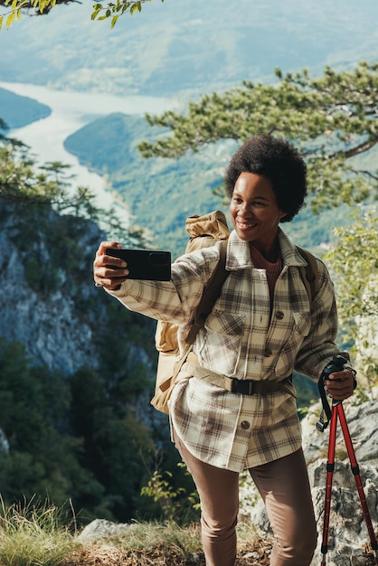 Mulher negra madura fazendo uma selfie em pé no topo de uma colina e apreciando a vista durante sua caminhada nas montanhas.