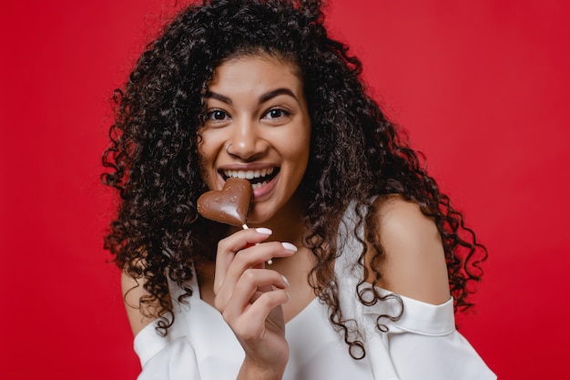 Foto mulher negra, mordendo o coração em forma de doces de chocolate e sorrindo na parede vermelha