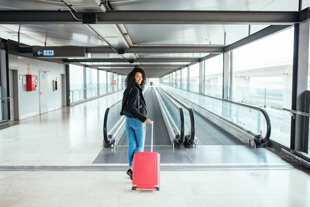 Mulher negra na passarela em movimento no aeroporto com uma mala rosa.