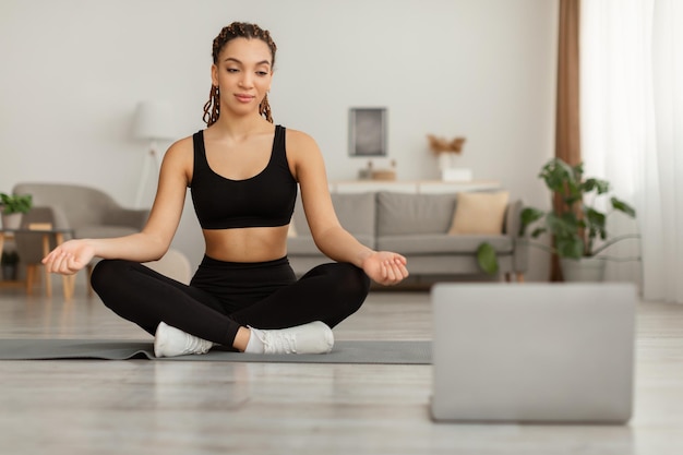 Mulher negra no laptop sentada em posição de lótus meditando dentro de casa