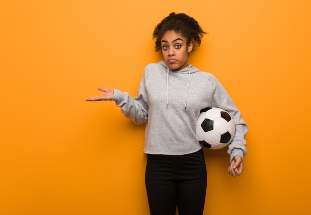 Mulher negra nova da aptidão que duvida e que shrugging ombros. Segurando uma bola de futebol.