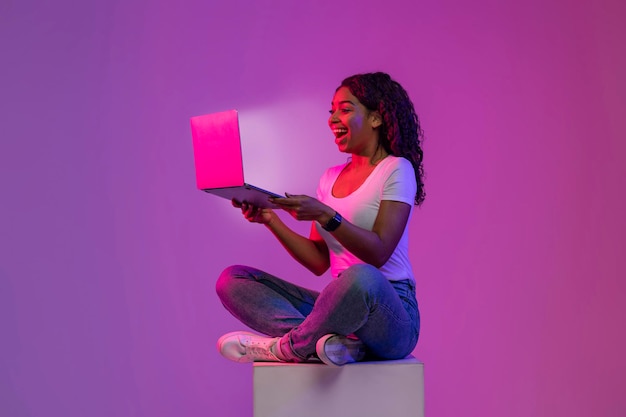 Mulher negra olhando para a tela brilhante do laptop enquanto está sentada no cubo grande