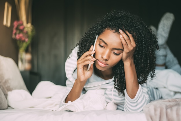 Mulher negra pensativa e triste séria bonita com cabelo encaracolado usando o smartphone na cama