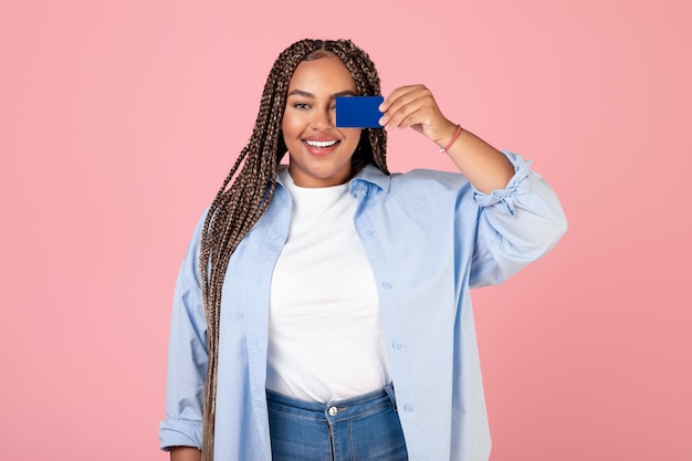 Mulher negra posando com cartão de crédito cobrindo o fundo rosa dos olhos