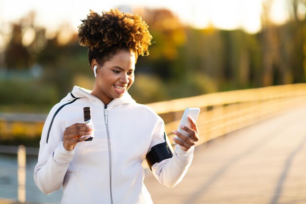 Mulher negra relaxando ao ar livre após o treino usando smartphone e comendo barra de proteína