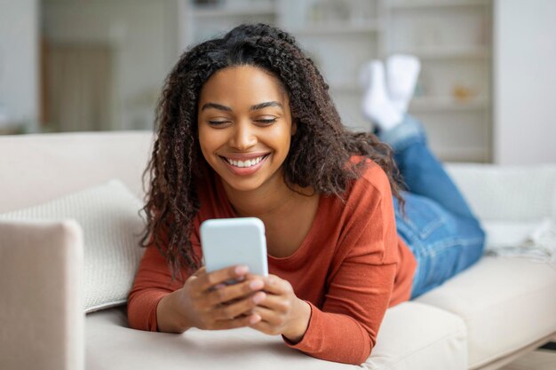 Mulher negra sorridente enviando mensagens de texto no smartphone enquanto relaxa em casa