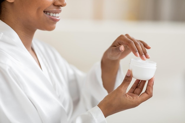 Mulher negra sorridente segurando o frasco aberto com creme hidratante fazendo tratamentos de beleza