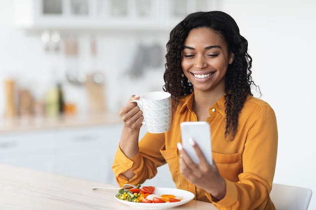 Mulher negra sorridente surfando nas mídias sociais enquanto toma café da manhã