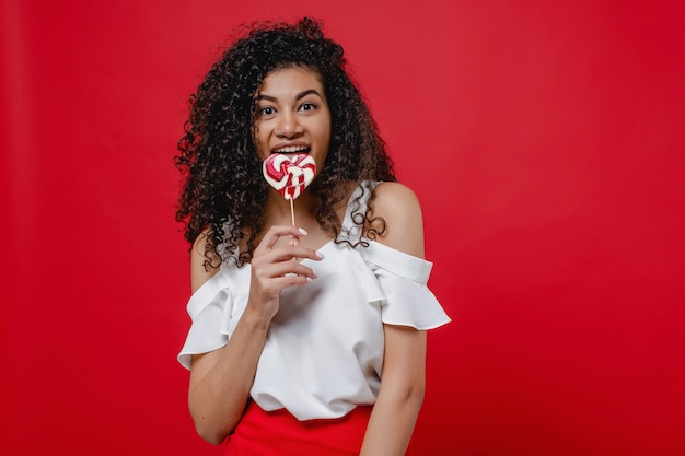 Mulher negra, sorrindo com doces pirulito em forma de coração isolados no vermelho