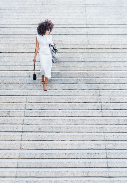 Mulher negra sozinha descendo as escadas olhando para o lado em um vestido branco