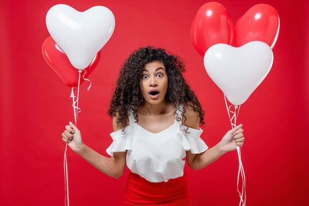 Mulher negra surpresa com coração colorido em forma de balões isolados no vermelho