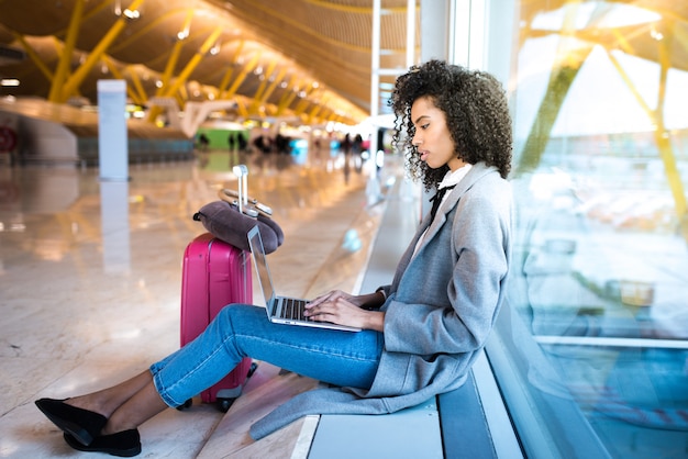 Mulher negra, trabalhando com o laptop no aeroporto esperando na janela