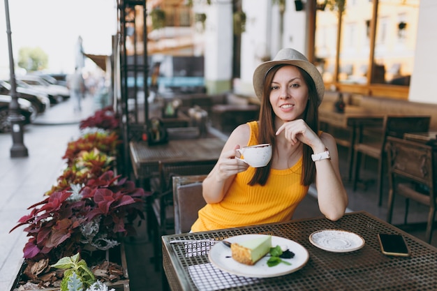 Mulher no café da rua ao ar livre, sentada à mesa, com um chapéu amarelo de roupas com uma xícara de cappuccino, bolo, relaxando no restaurante durante o tempo livre