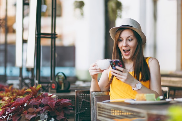 Mulher no café da rua ao ar livre, sentada à mesa com um chapéu com uma xícara de bolo de cappuccino, usando o telefone celular, relaxando no restaurante no tempo livre