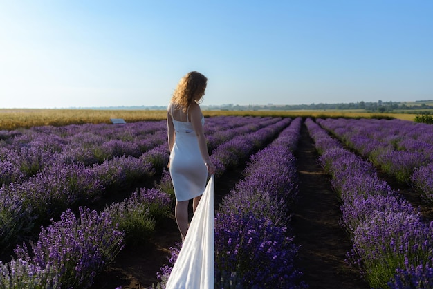 Mulher no campo de flores de lavanda ao pôr do sol em vestido branco andando de manhã