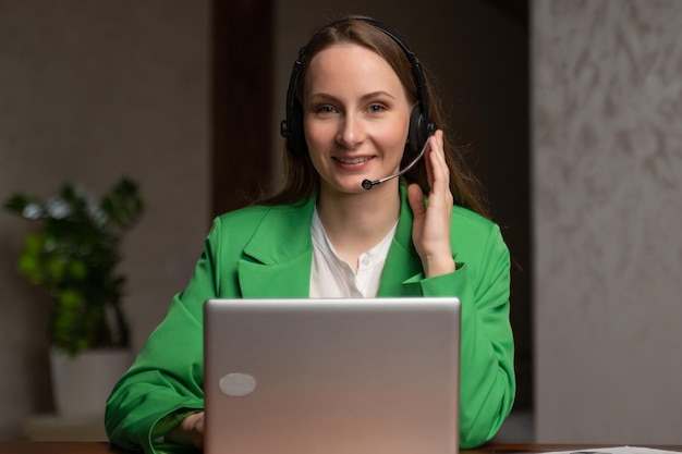 Mulher no fone de ouvido fala com o cliente sentado perto do laptop aberto à mesa no escritório jovem consultora feminina