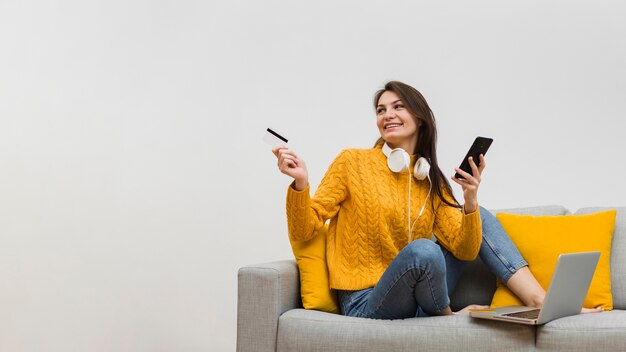 Mulher no sofá, segurando o smartphone em uma mão e um cartão de crédito na outra