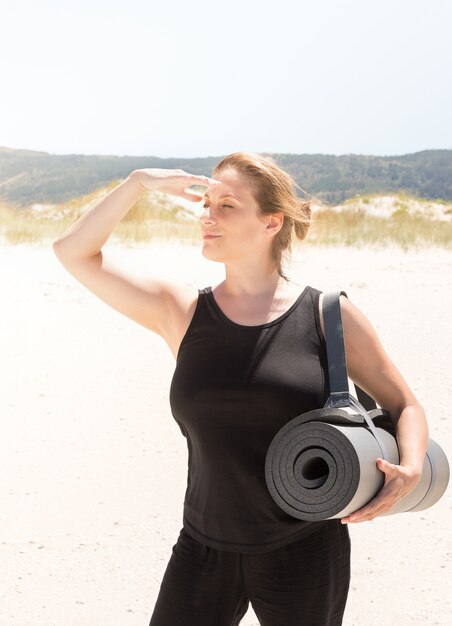 Mulher no sportswear com um tapete enrolado debaixo do braço na praia