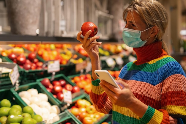 Mulher no supermercado na seção de frutas escolhendo frutas com smartphone usando uma máscara de proteção antivírus devido à pandemia de coronavírus covid19