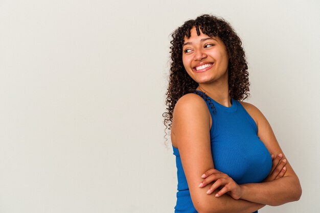 Mulher nova da raça misturada isolada no fundo branco, sorrindo confiante com os braços cruzados.