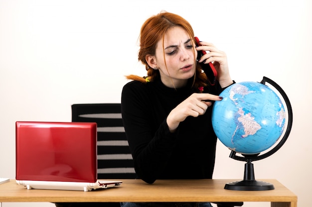 Mulher nova do agente de viagens que senta-se atrás da mesa de trabalho com laptop e globo geográfico do mundo que fala em um telefone celular.