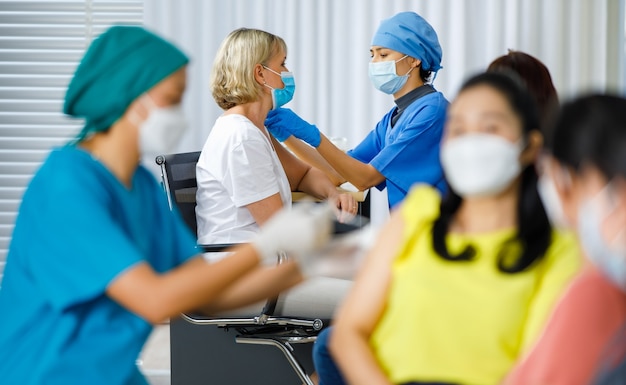 Mulher octor usa máscara facial, luvas de borracha e uniforme azul do hospital, injetando vacina em uma mulher caucasiana, enquanto outra enfermeira vacina uma paciente na fila na clínica em primeiro plano.