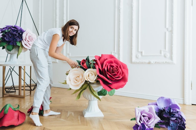 Mulher ocupada vestida casualmente decora salão com flores para evento festivo Florista feminina prepara salão de banquetes para casamento usa diferentes composições de flores